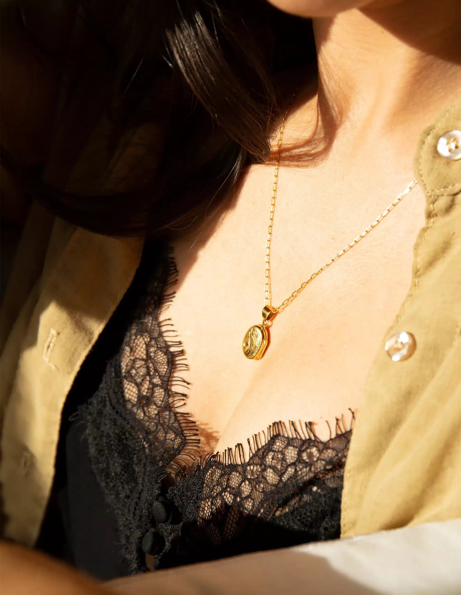 En kvindes hals og bryst er synlige. En guld halskæde med en medaljon hænger over en sort blonde bh og en åben beige skjorte.