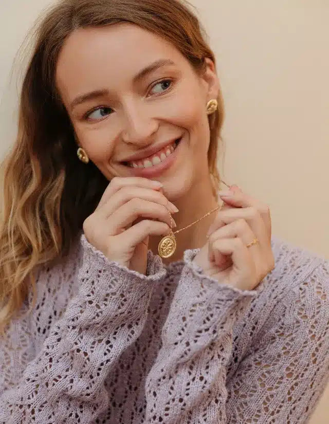 En smilende kvinde med blondt hår holder en guldfarvet halskæde med et rundt vedhæng, iført en lilla striktrøje, foran en beige baggrund.