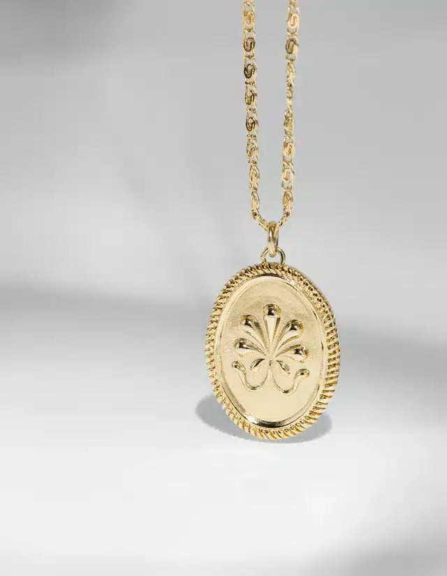 En gylden halskæde med en oval vedhæng hænger ned. Vedhænget har et blomsterlignende design indgraveret og en snoet kant, der giver ekstra detaljer. Det hele ses mod en lys, neutral baggrund.