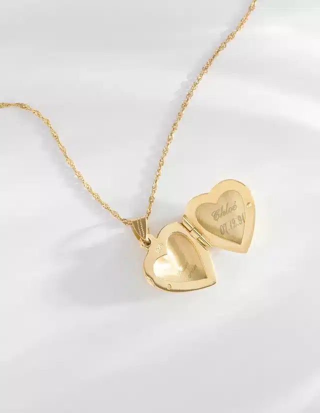 En guldhalskæde ligger på en hvid baggrund, med et åbent hjerteformet vedhæng; vedhænget er indgraveret med "Chloe 07.19.91".