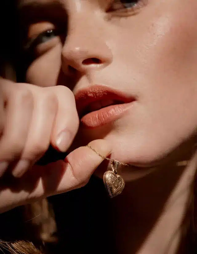 En kvindes hånd holder i en guldhalskæde med et hjerteformet vedhæng, mens hendes fingre let rører ved hendes læber. Det hele er fotograferet i et intimt, varmt lys.