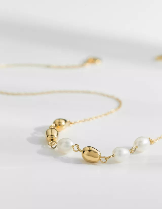 En guldkæde er udsmykket med perler og guldperler, der ligger på en hvid overflade. Kæden snor sig let og reflekterer lyset.