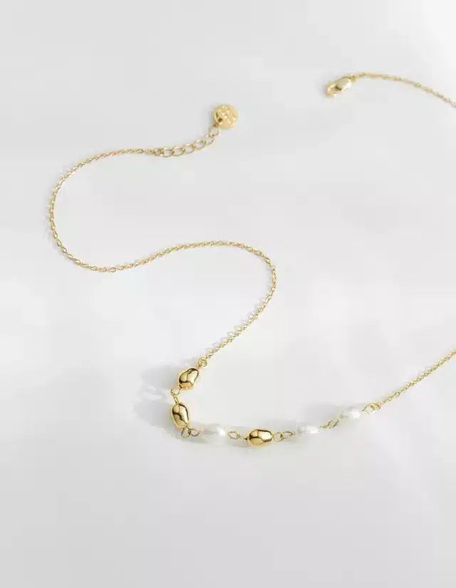 En gylden halskæde ligger på en hvid overflade. Den har små guldperler og perler spredt langs kæden.