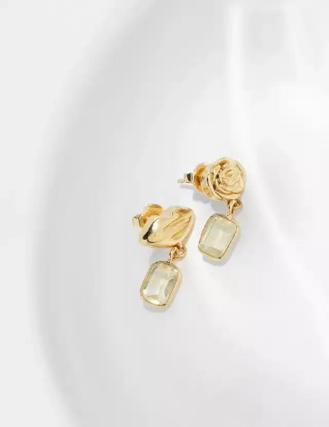 Guldøreringe med en rosetformet top hænger over en hvid baggrund. En facetslebet rektangulær sten hænger fra hver ørering.