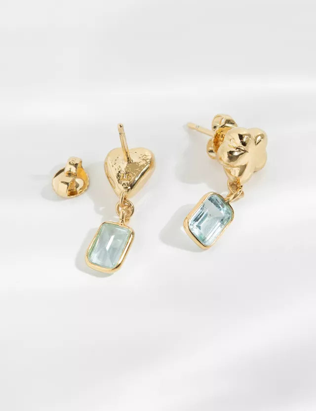 Guldøreringe med lyseblå rektangulære vedhæng ligger på en hvid overflade. Den ene er en hjerteformet ørestik, den anden er formet som et firkløver.