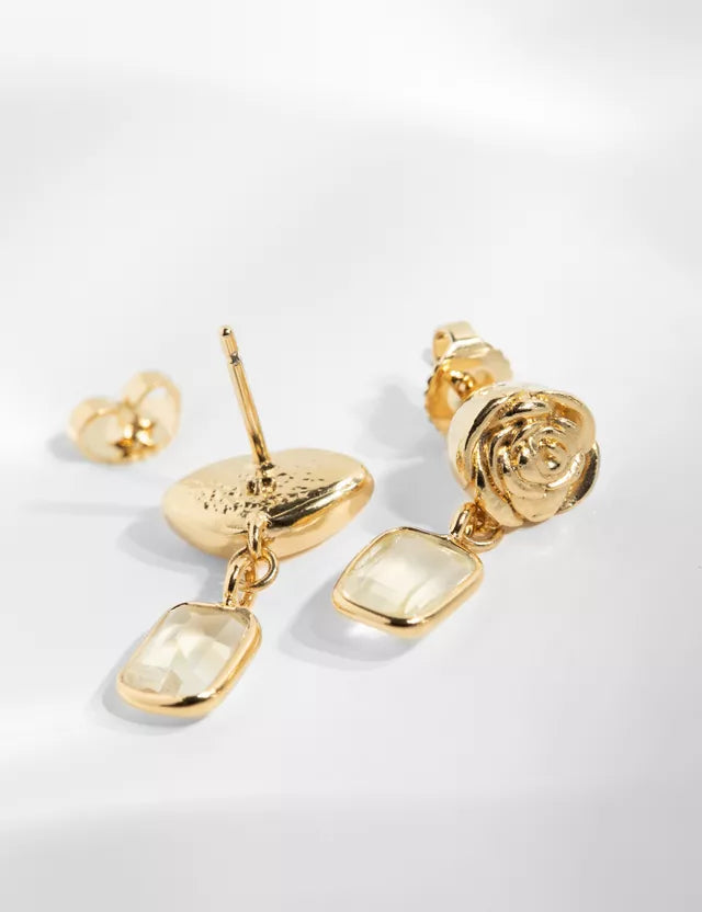 Guldøreringe med en rose og en rektangulær krystal hænger over en hvid overflade, sammen med øreprops.