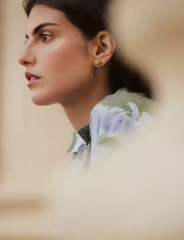 En kvinde med mørkt hår vises i profil, iført en blomstret skjorte og rektangulære øreringe, i et blødt, diffust miljø.