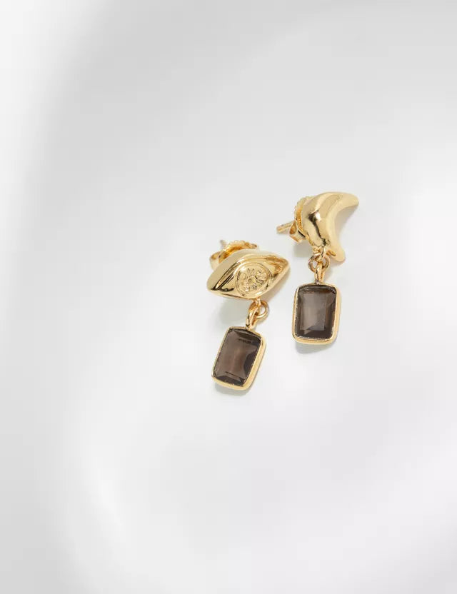 To gyldne øreringe, en med et øje og en med en bøjet form, hænger på en hvid overflade. Hver ørering har en rektangulær, brunlig sten hængende nedenunder.