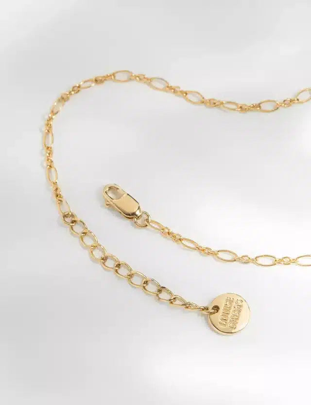 En guldkæde ligger på en hvid overflade. Den har en hummerlås og en lille rund disk med teksten "LOUISE DAMAS."