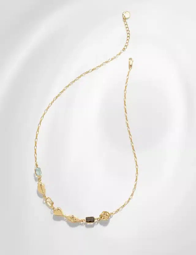 En guldkæde ligger mod en hvid baggrund. Kæden er pyntet med forskellige ædelsten i forskellige former, herunder en rose.