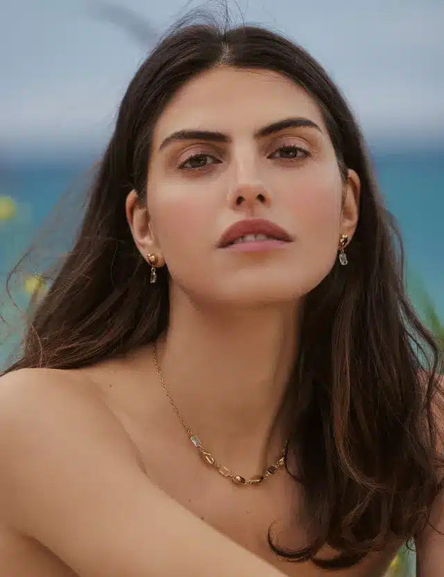 En kvinde med mørkt hår og smykker ser direkte på beskueren. Hun er udendørs med en baggrund af hav og vegetation.