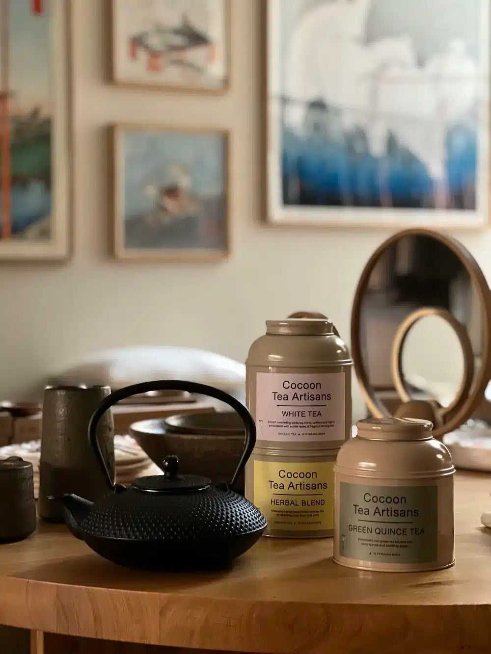 En sort tekande sidder på et træbord sammen med "Cocoon Tea Artisans" tebeholdere: "WHITE TEA", "HERBAL BLEND" og "GREEN QUINCE TEA". Kunst hænger på væggen bagved.