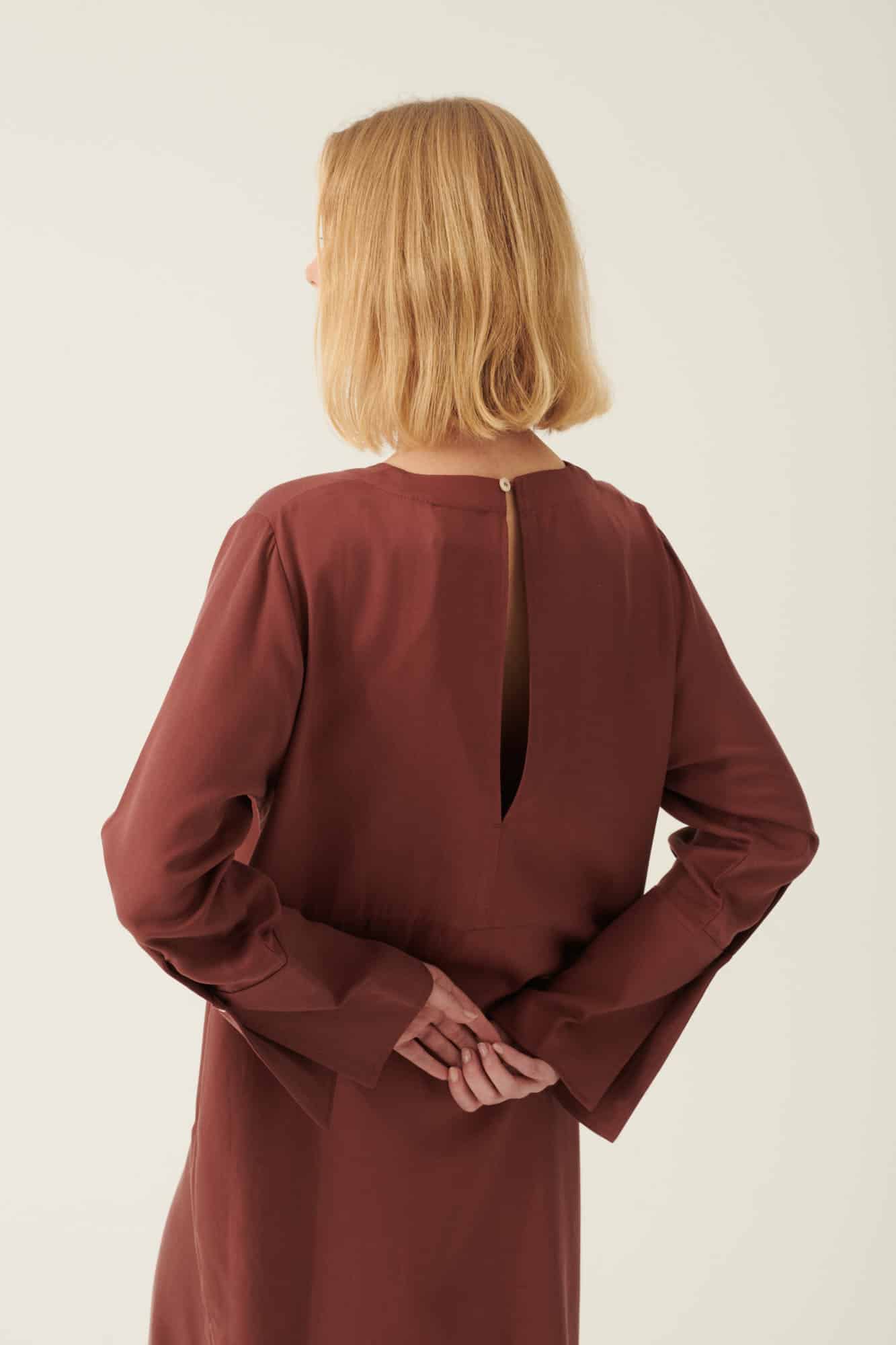 En kvinde med blond pagefrisure står med hænderne foldet bag ryggen iført en vinrød kjole med lange ærmer og en lille åbning i nakken. Baggrunden er cremefarvet.