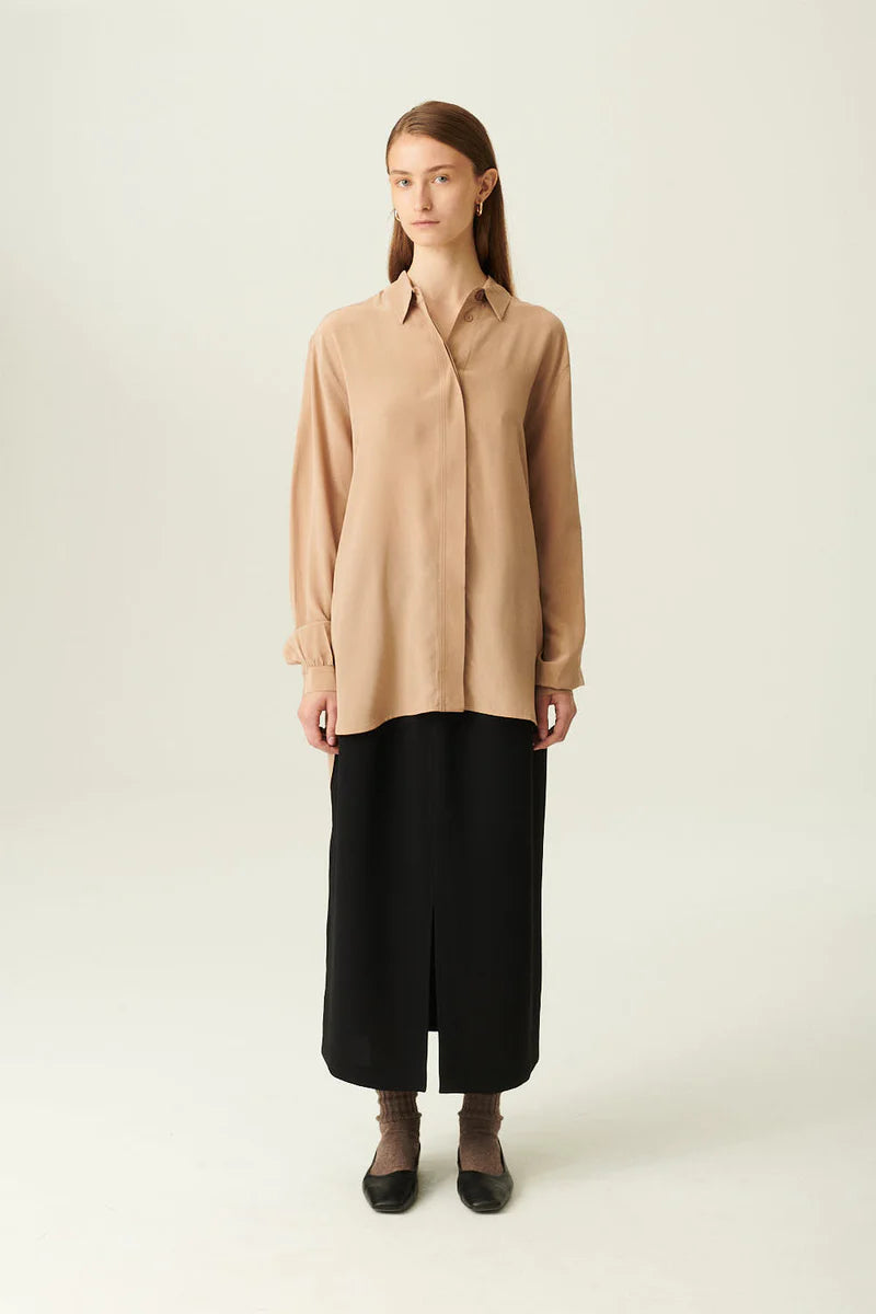 En kvinde står i et lyst studie med en beige, langærmet skjorte og en sort, lang nederdel med slids. Hun har sokker og sorte sko på.