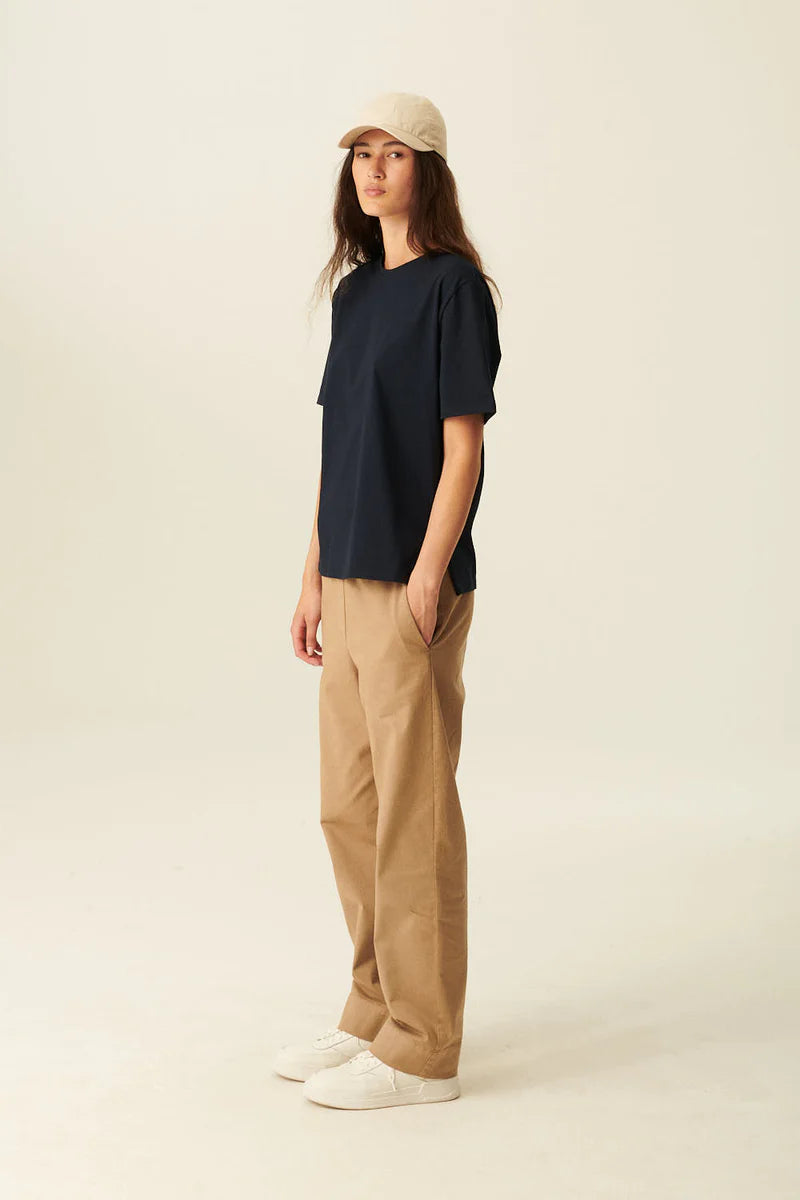 En kvinde poserer i et studie, iført en mørkeblå t-shirt, kaki bukser, hvide sneakers og en beige kasket. Hun har en hånd i lommen.