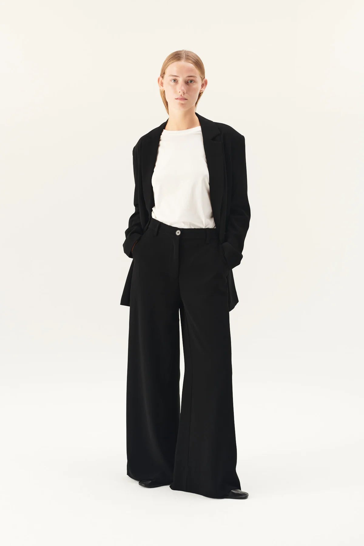 En kvinde står med hænderne i lommen på et sort jakkesæt med vide ben. Hun har en hvid t-shirt på under jakken, og baggrunden er hvid.