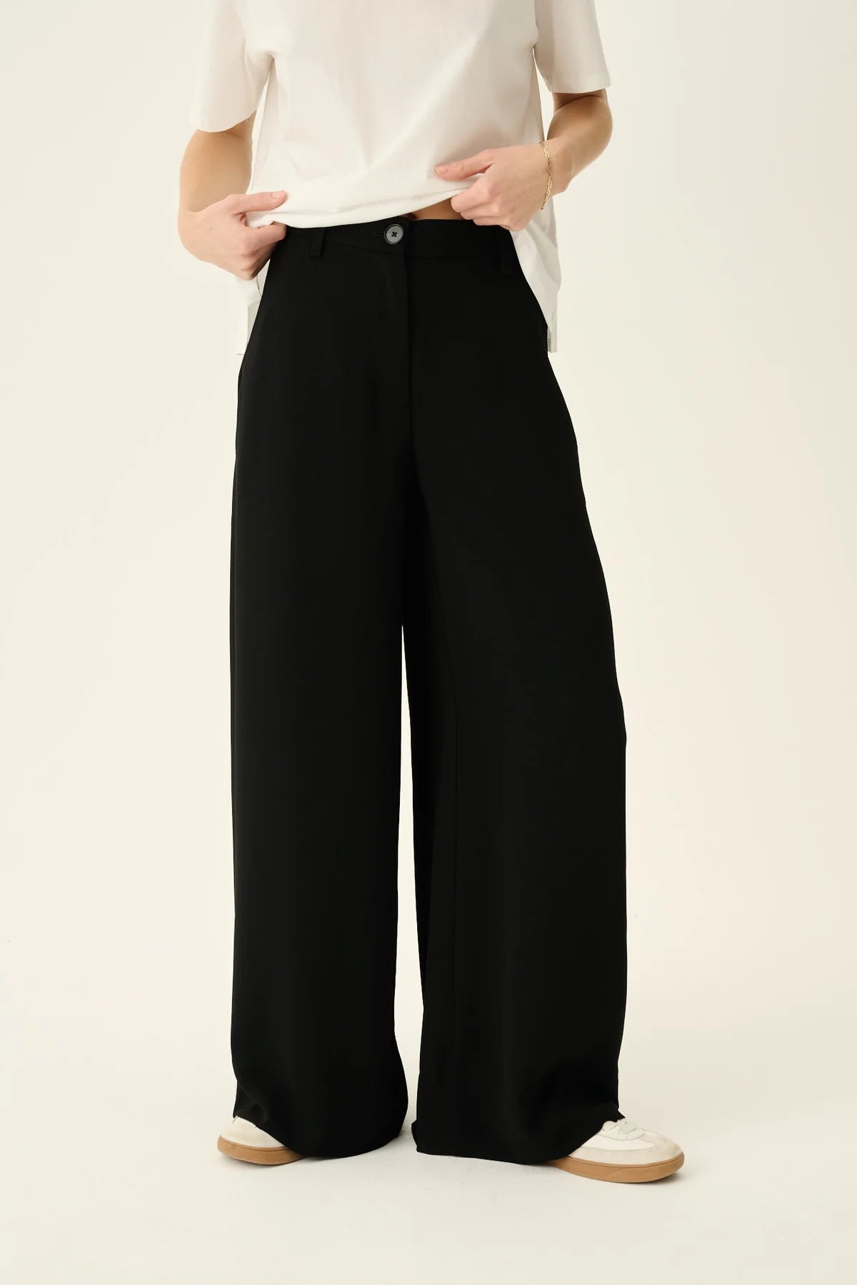 En person iført en hvid T-shirt og vide, sorte bukser står i et studiemiljø med en cremefarvet baggrund. Hænderne holder nederst i T-shirten.