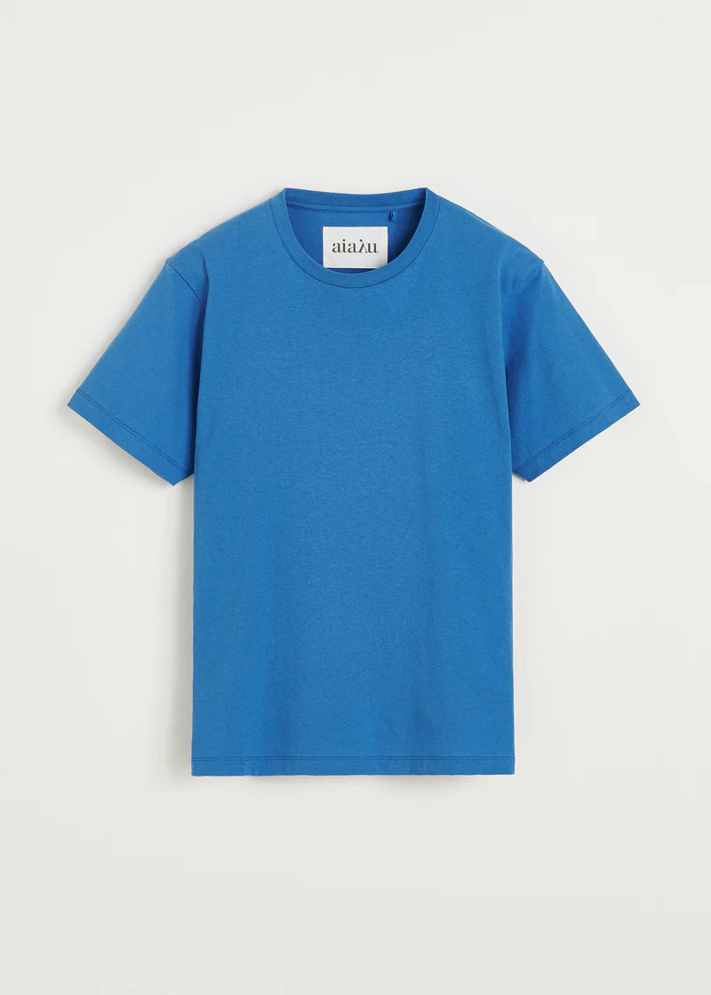 En lyseblå T-shirt ligger fladt på en hvid baggrund. T-shirten har et "aiaľu" mærke synligt i nakken.