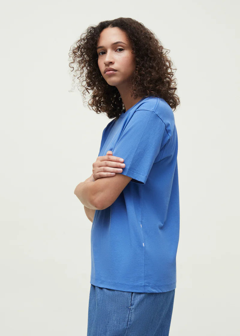 En kvinde med krøllet hår poserer iført en blå T-shirt og denim shorts. Hun står foran en lys baggrund med armene krydset.