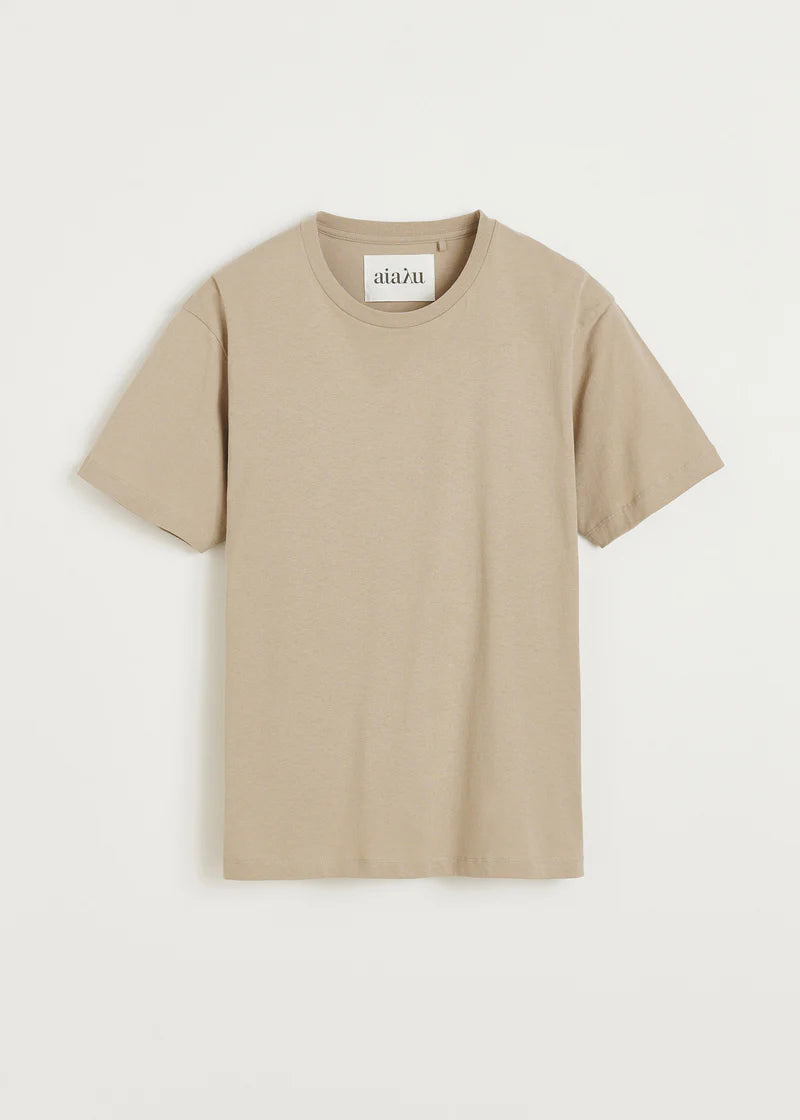 En beige t-shirt med korte ærmer og mærket "aiaλu" er placeret centreret på en hvid baggrund.