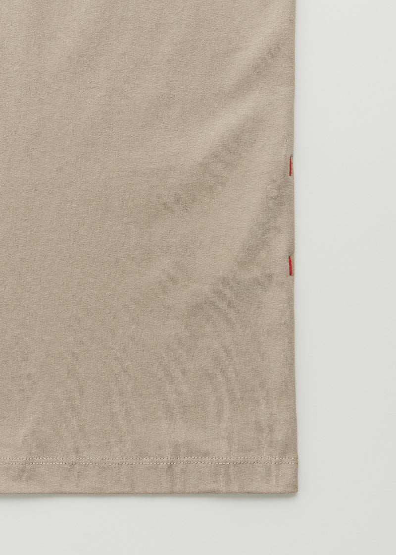 En beige t-shirt viser sin nederste kant og sidesøm mod en hvid baggrund. Sidesømmen er fastgjort med røde sting.