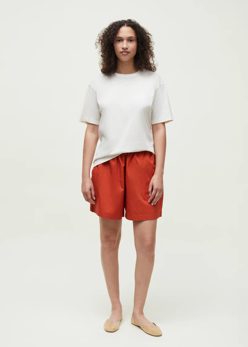 En kvinde står, iført en hvid t-shirt og orange shorts, på en ensfarvet, hvid baggrund. Hun har krøllet hår og beige sko.