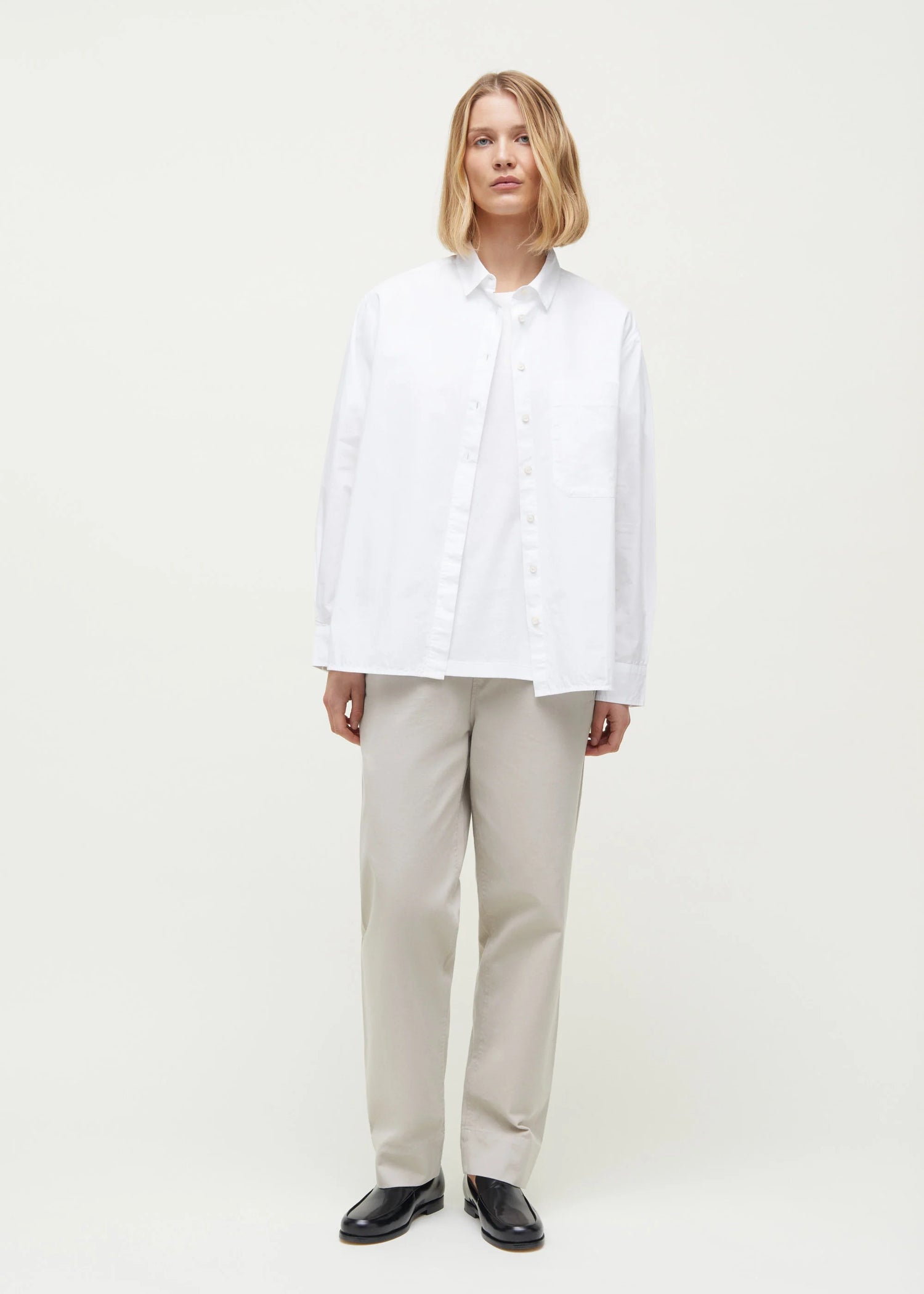 En kvinde står i et hvidt miljø og viser tøj: en hvid skjorte over en hvid T-shirt, beige bukser og sorte sko.
