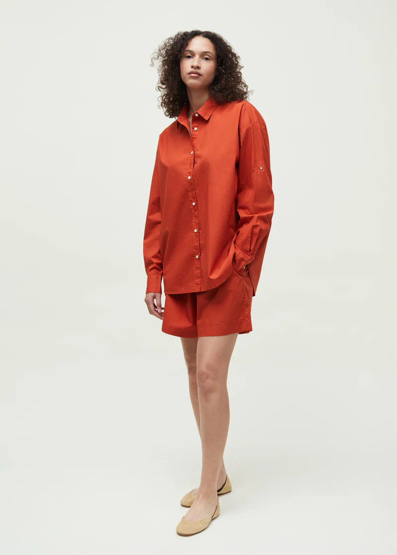 En kvinde med krøllet hår står, iført en orange skjorte og shorts, med den ene hånd i lommen, på en hvid baggrund. Hun har beige sko på.