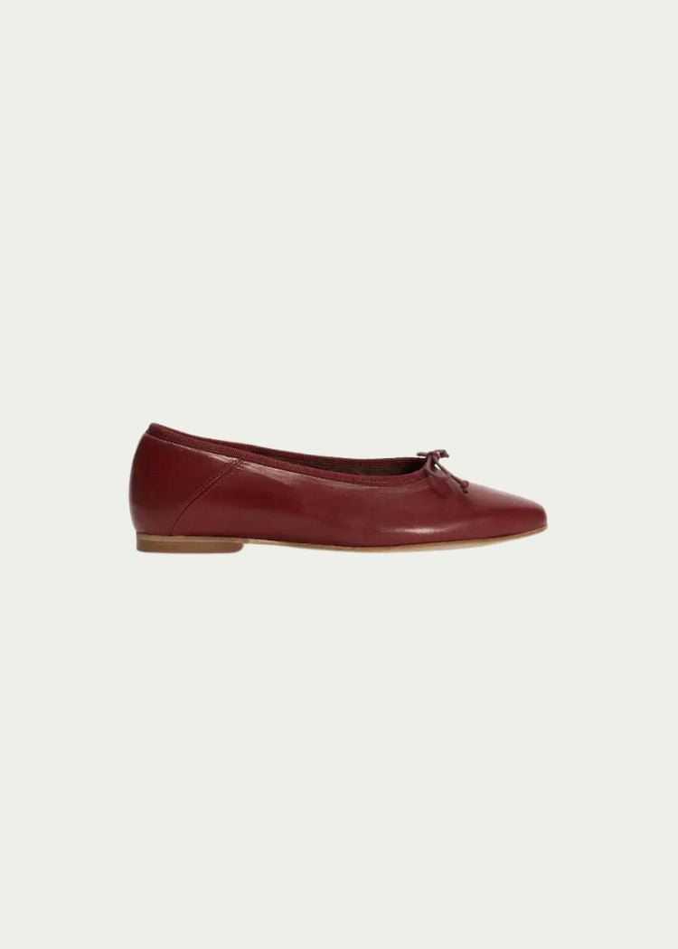 En rød, læder ballerina sko hviler, stillestående, mod en neutral, ensfarvet baggrund. Skoen har en lille sløjfe på tåen.