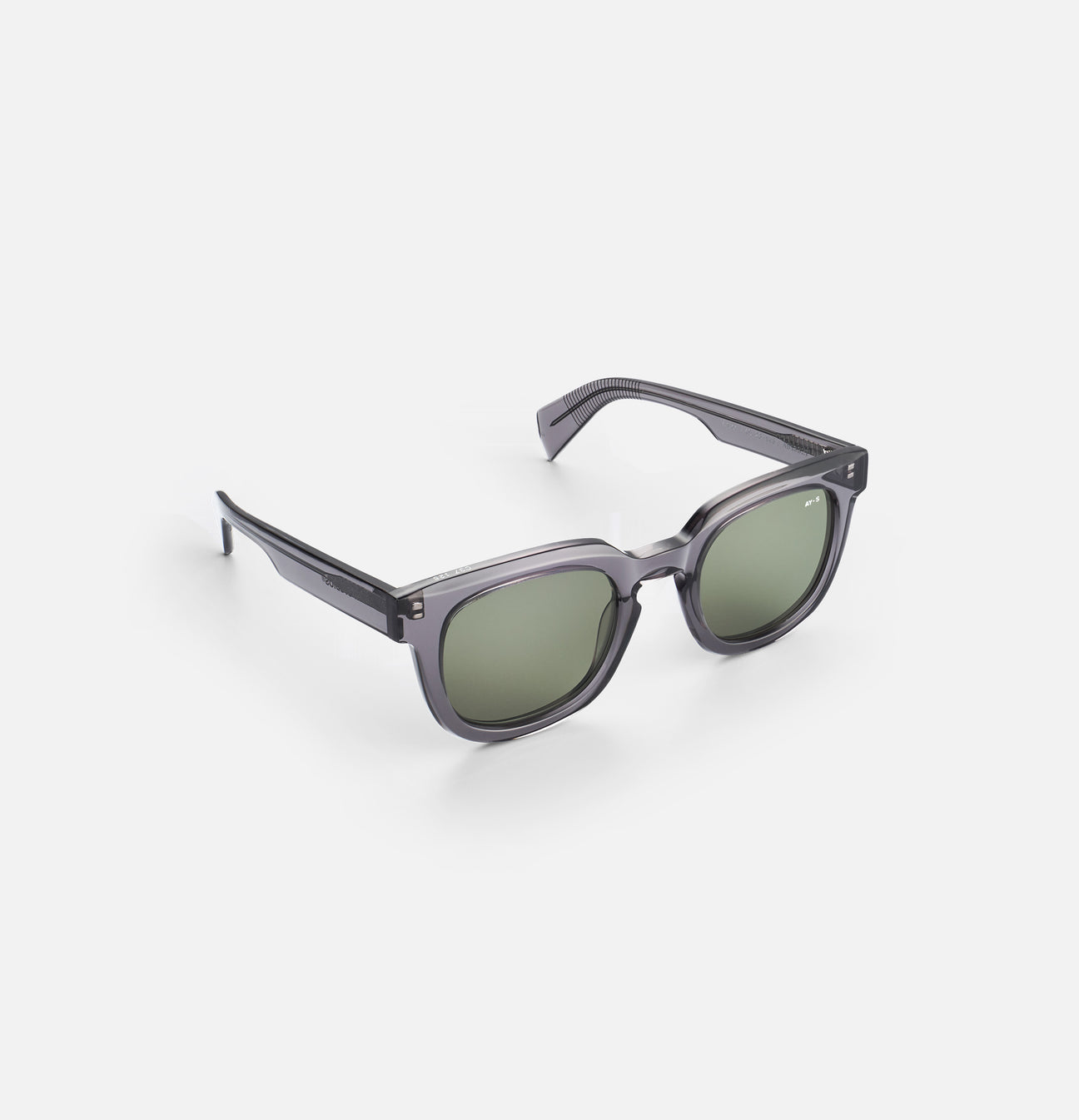 Grå solbriller med grønne linser og firkantet ramme ligger skråt på en hvid overflade. "AY-S" er skrevet på højre side af rammen.