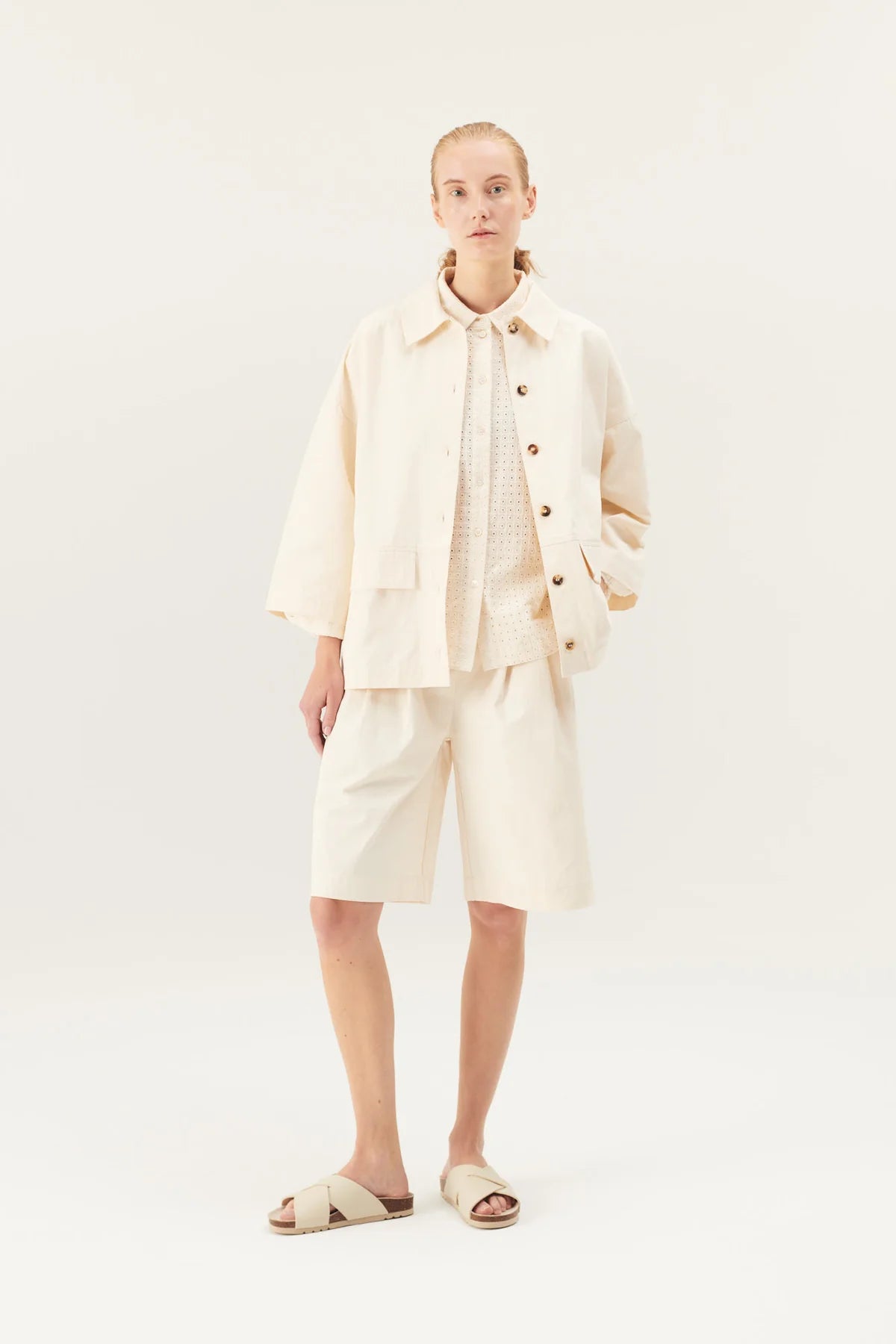 En kvinde står i et studiemiljø. Hun er iført en matchende beige jakke og shorts over en skjorte og sandaler.