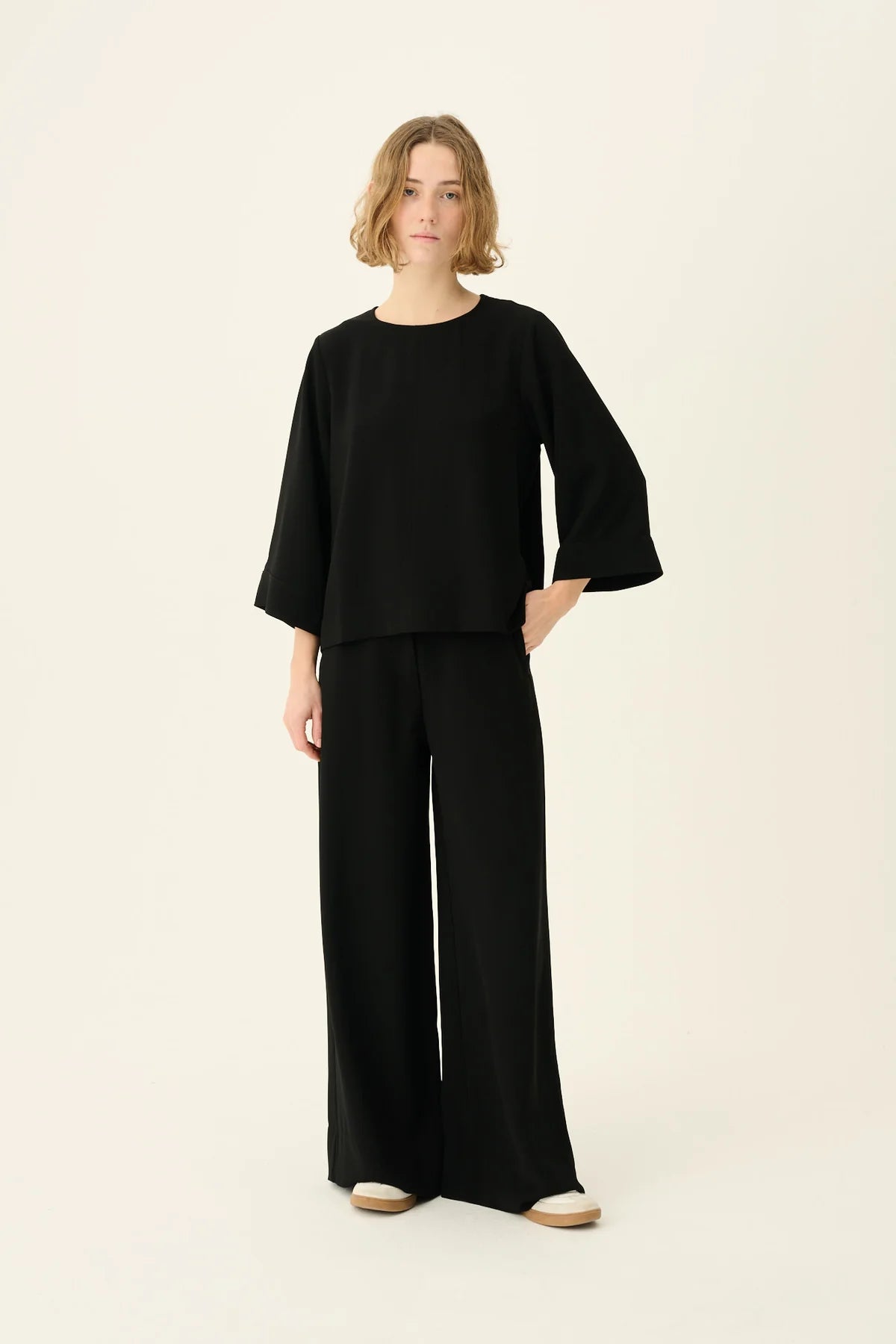 En kvinde i sort tøj står mod en ensfarvet baggrund. Hun har en hånd i lommen og kigger direkte på kameraet.