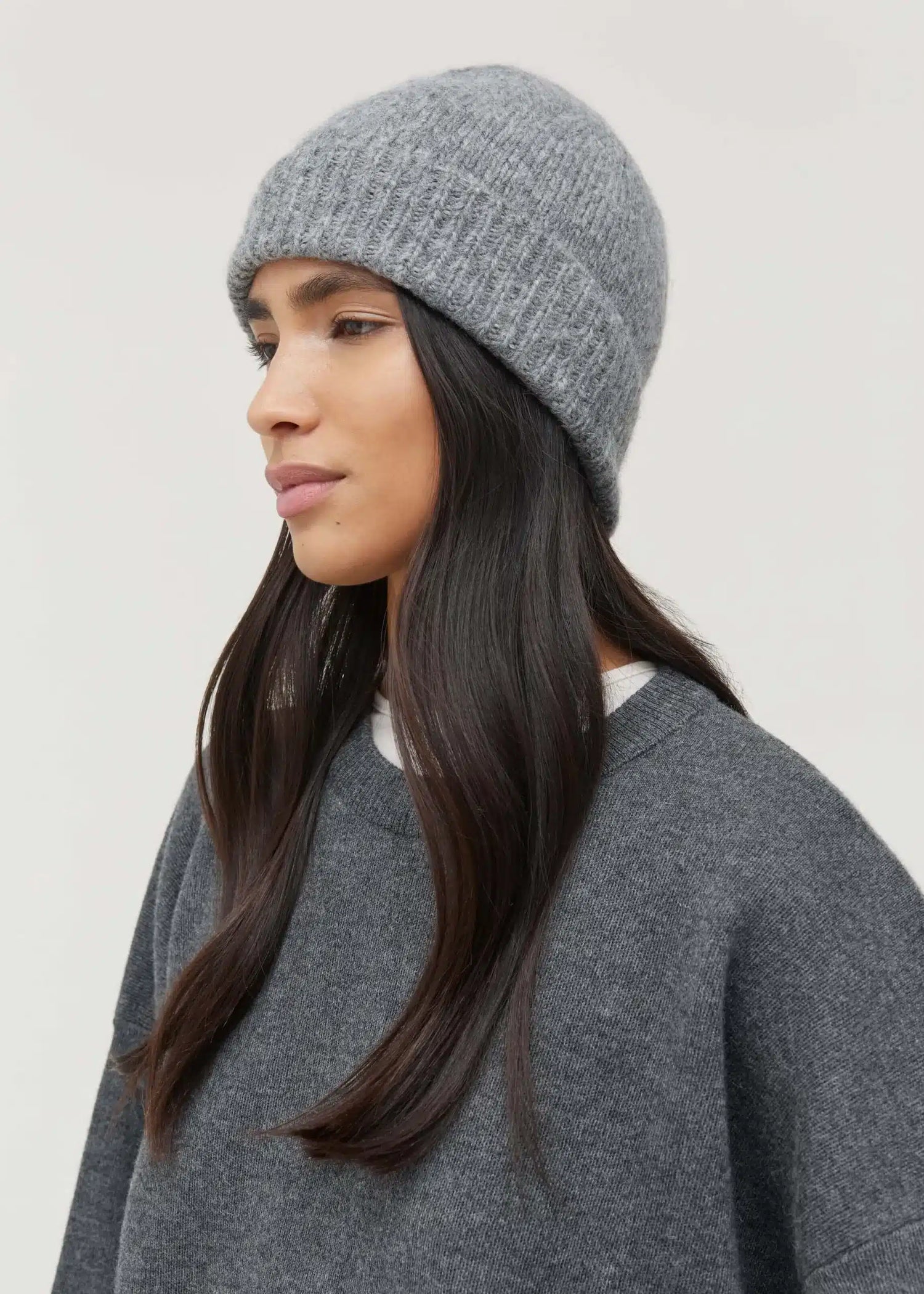 En kvinde bærer en grå strikhue og en grå sweater i en neutral baggrund. Kvinden kigger til venstre.