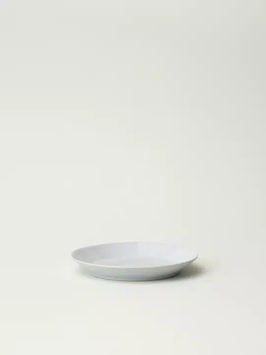 En hvid tallerken hviler på en lys, ensfarvet baggrund. Tallerkenen er rund og ser ud til at være tom.