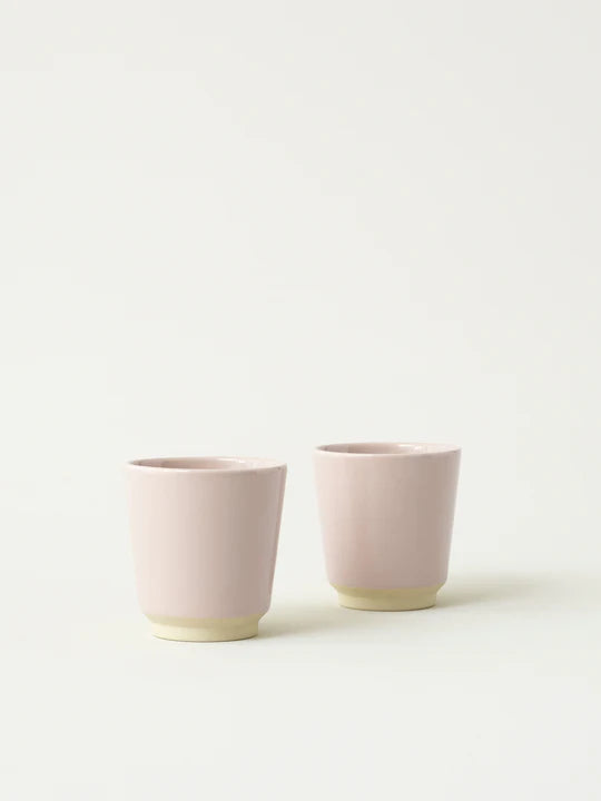 To lyserøde keramikkopper står stille ved siden af hinanden på en ensfarvet, off-white baggrund. Kopperne har en beige base.