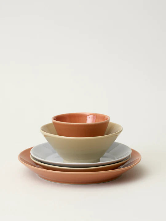 En stak af forskellige farvede keramikskåle og tallerkener er stablet oven på hinanden på en off-white baggrund. Farverne er primært jordfarver.