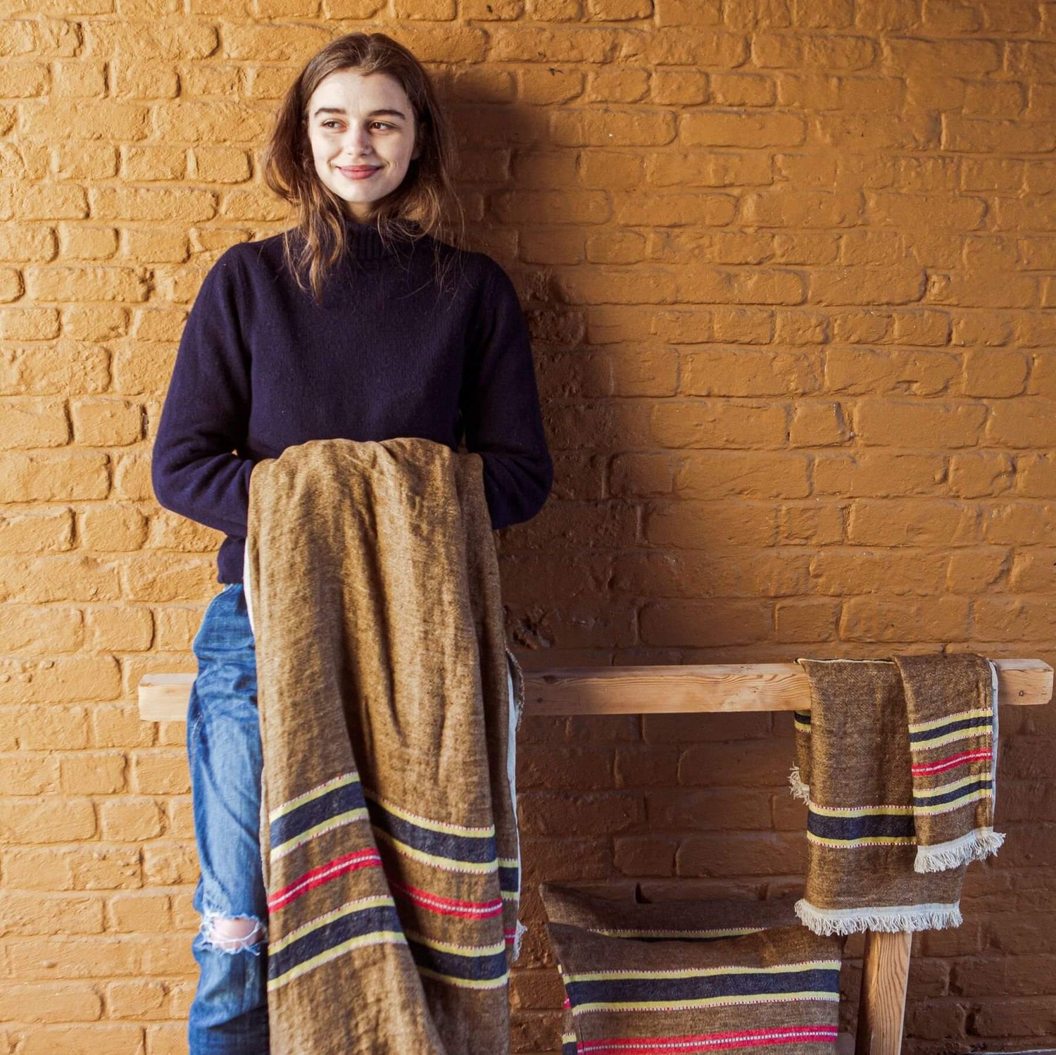 En kvinde holder et stribet tæppe foran en murstensvæg. Et lignende tæppe hænger over en træbænk, og en pude ligger under den.
