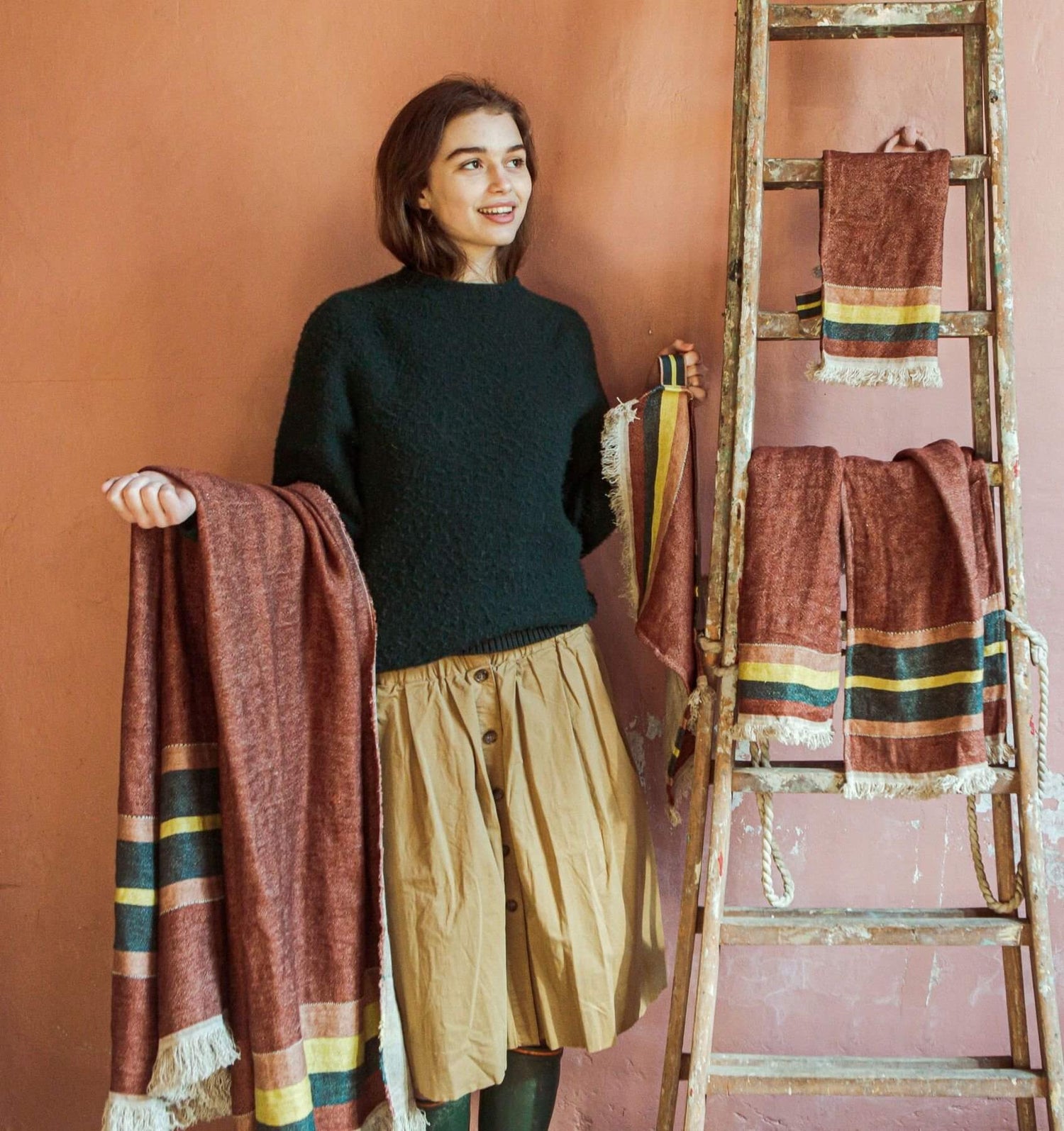 En kvinde holder et brunligt tæppe med farvede striber og frynser foran en rosa væg. Ved siden af står en gammel stige med flere af tæpperne hængende over trinnene.