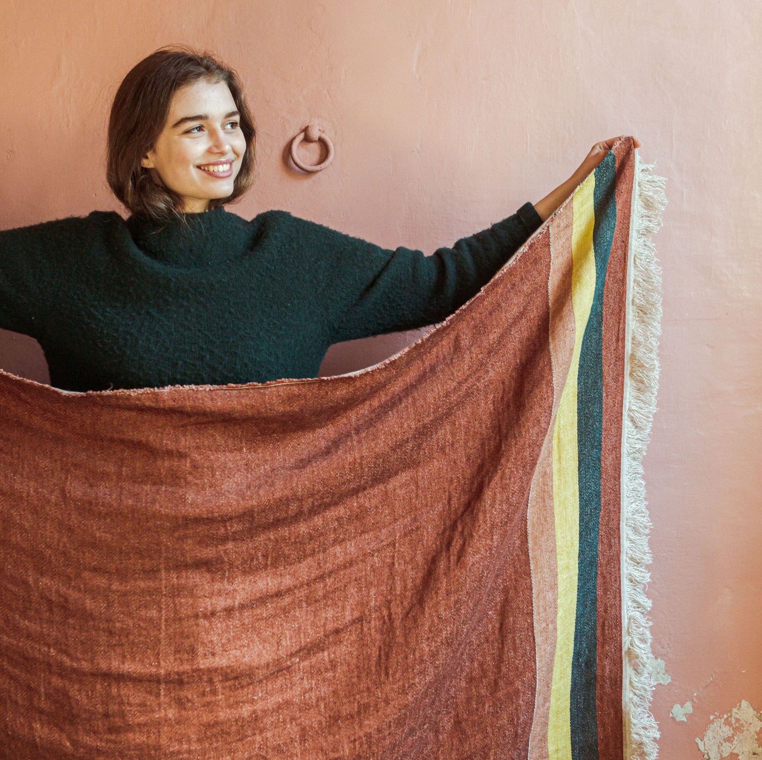 En smilende kvinde i en grøn sweater holder et brunt tæppe med farvede striber og frynser foran en lyserød væg.