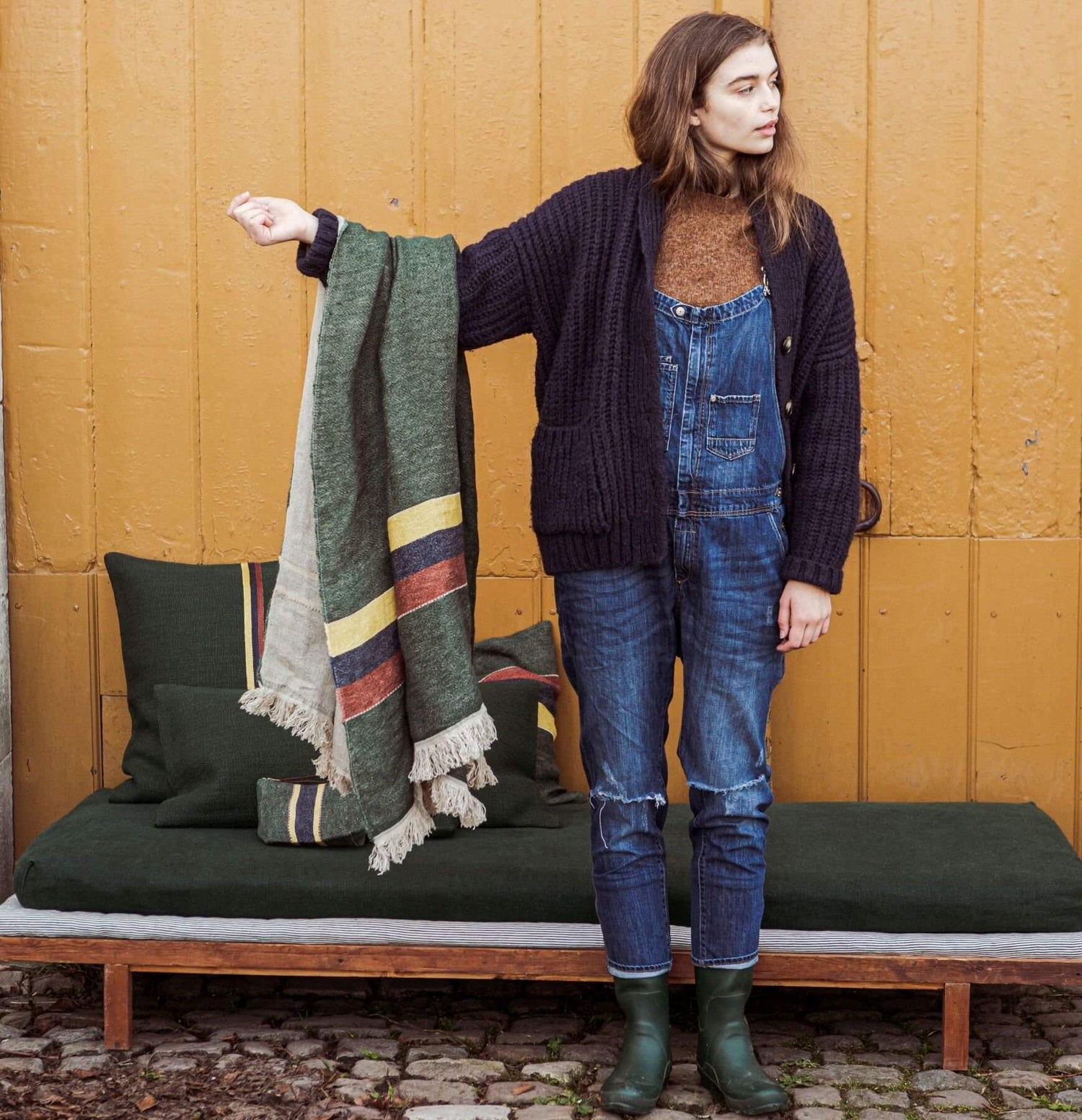 En kvinde holder et tæppe med striber, ved siden af en bænk med puder og baggrund af en gul væg. Hun er iført overalls og gummistøvler.