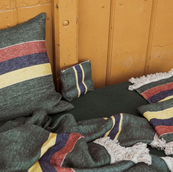 Grønne tekstiler med striber i rød, gul og blå ligger mod en gul trævæg. En pude, et tæppe, en lille taske og en dekoration er vist.