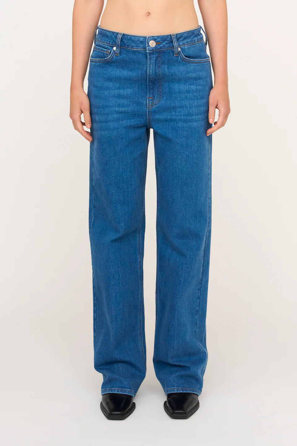En person iført blå jeans står på en hvid baggrund. Bukserne har en lige pasform, og der er sorte sko synlige under buksernes kant.