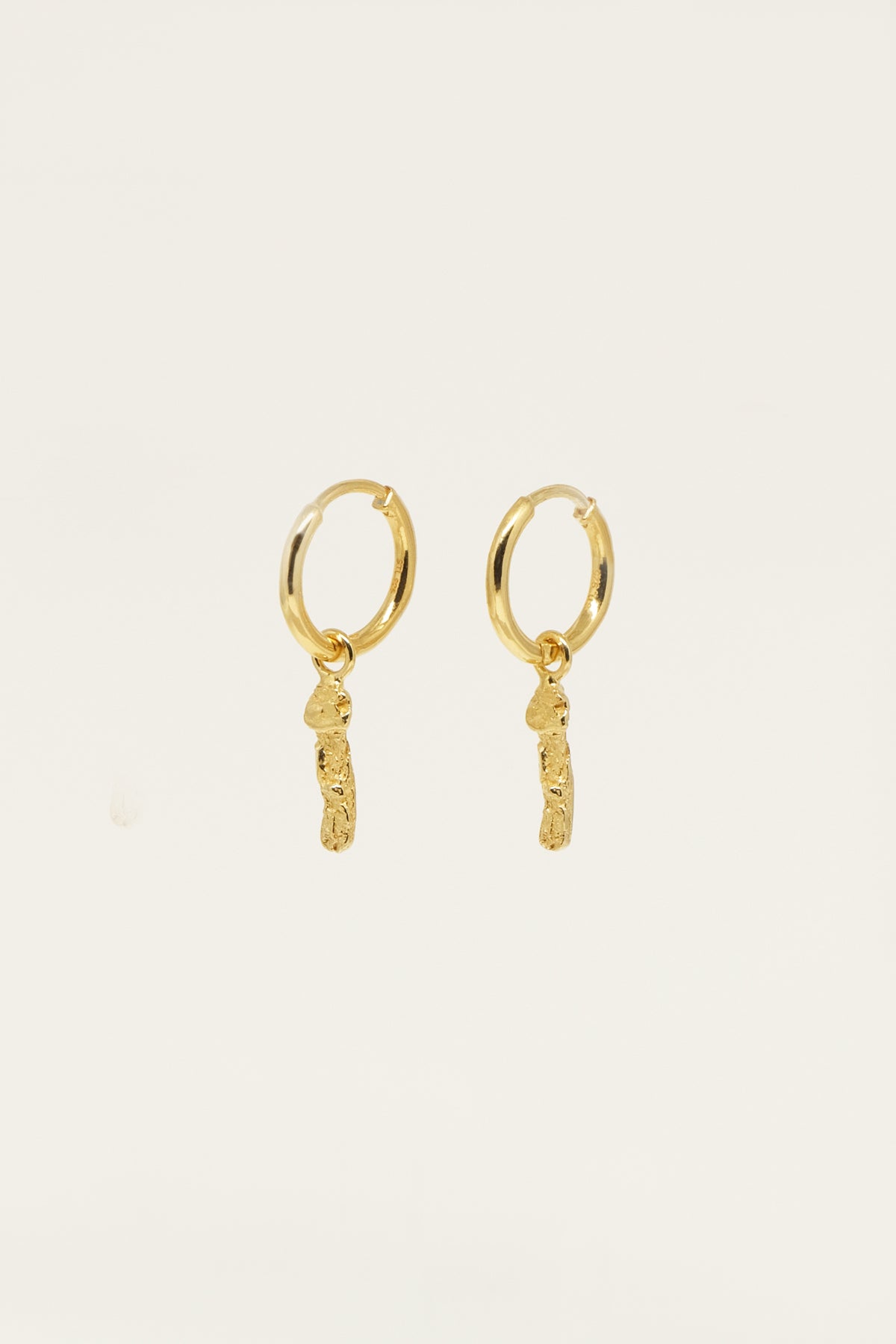 To gyldne hoop-øreringe hænger mod en neutral, off-white baggrund. Fra hver hoop hænger et lille, struktureret, guldfarvet vedhæng.