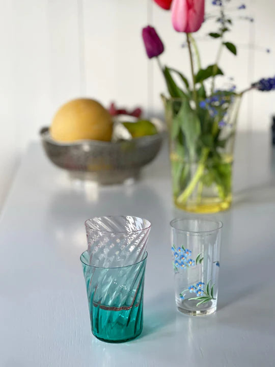 Glas ses på et bord; to er stablet, en anden har blomster malet på den. En skål med frugt og en vase med tulipaner er i baggrunden.