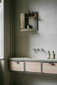 Et badeværelsesmiljø med en vaskekommode i træ og metal, over hvilken der er monteret en metalhylde med diverse flasker og glas. Armaturet er af metal med krydsformede håndtag.