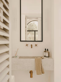 En vask af sten står i et hvidt badeværelse, med et sort indrammet spejl og guld vandhane. Et håndklæde hænger over vasken, og to flasker står på siden.