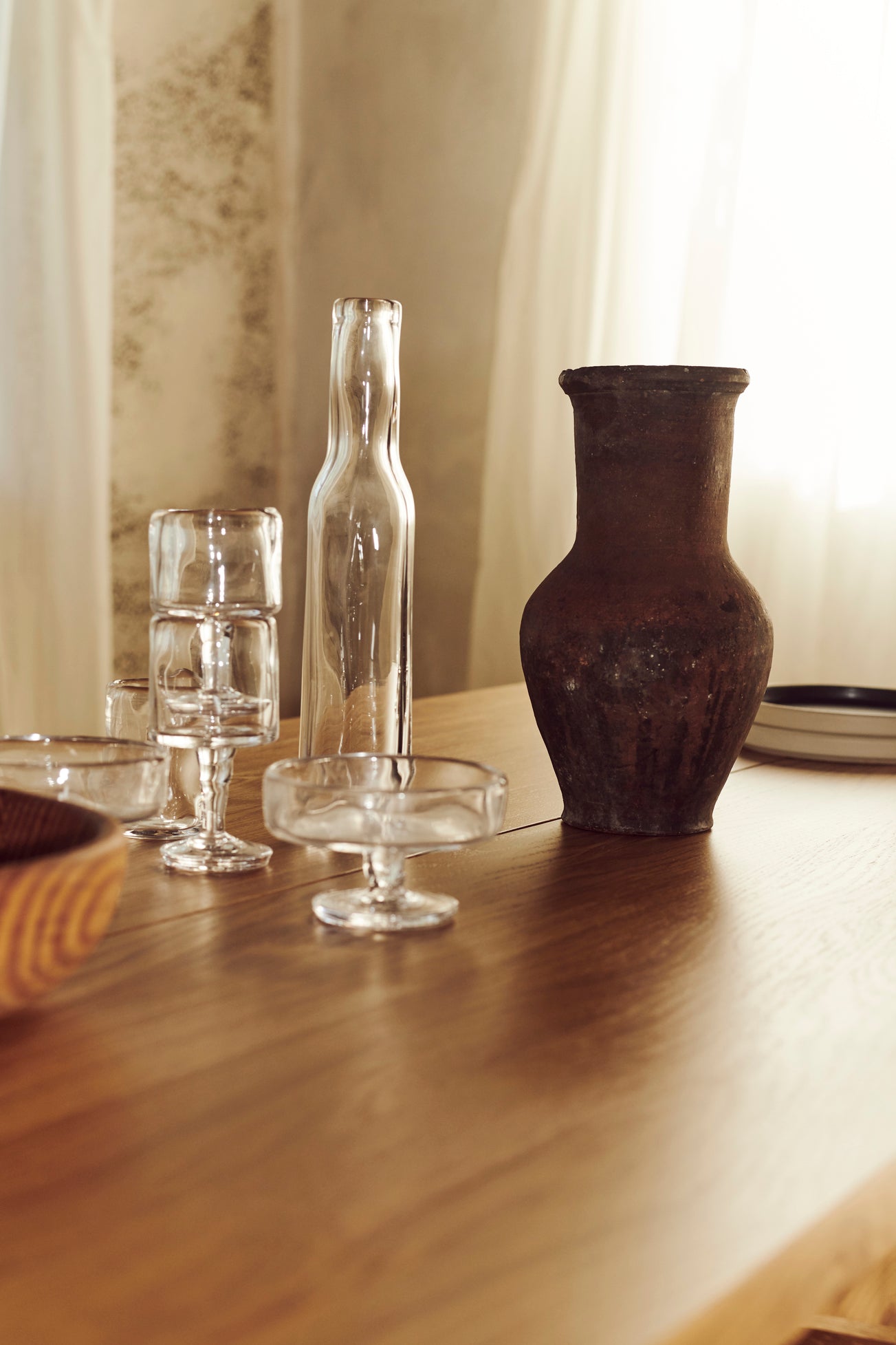 Glasvarer og en mørk krukke står stille på et lyst træbord. En flaske og stablede glas ligger til venstre for krukken, med en dæmpet baggrund.