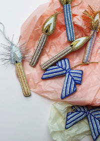 Beadsformede juleornamenter, inklusiv lys og sløjfer, ligger på et baggrundsmateriale af blød, krøllet papir. Ornamenterne har guld eller sølv tråd til ophæng.