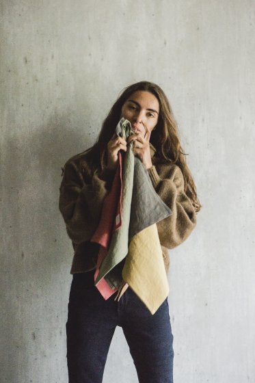 En kvinde holder farverige klude op til sit ansigt. Hun står foran en grå betonvæg og er iført en brun sweater og mørke jeans.