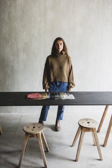 En kvinde står foran et langt, mørkt bord. Hun betragter stofprøver på bordet, i et lyst rum med minimalistisk indretning og træskamler.
